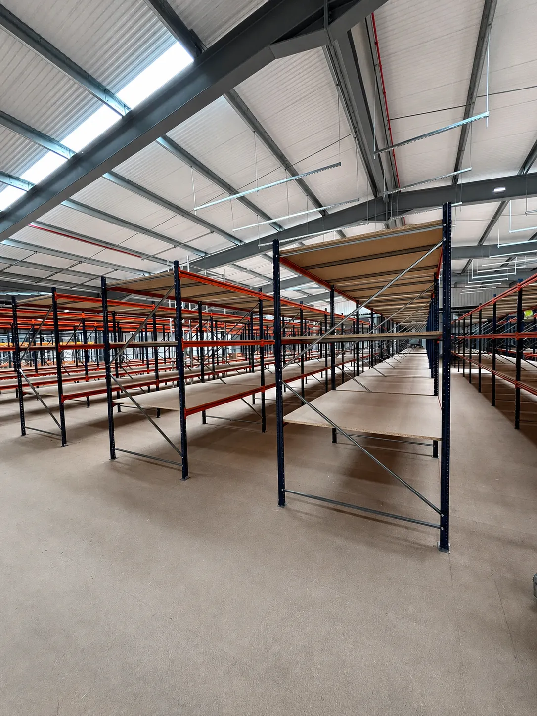 Runs of newly installed long-span shelving with braced frames and timber shelf levels, either side of wide aisles in a warehouse unit.